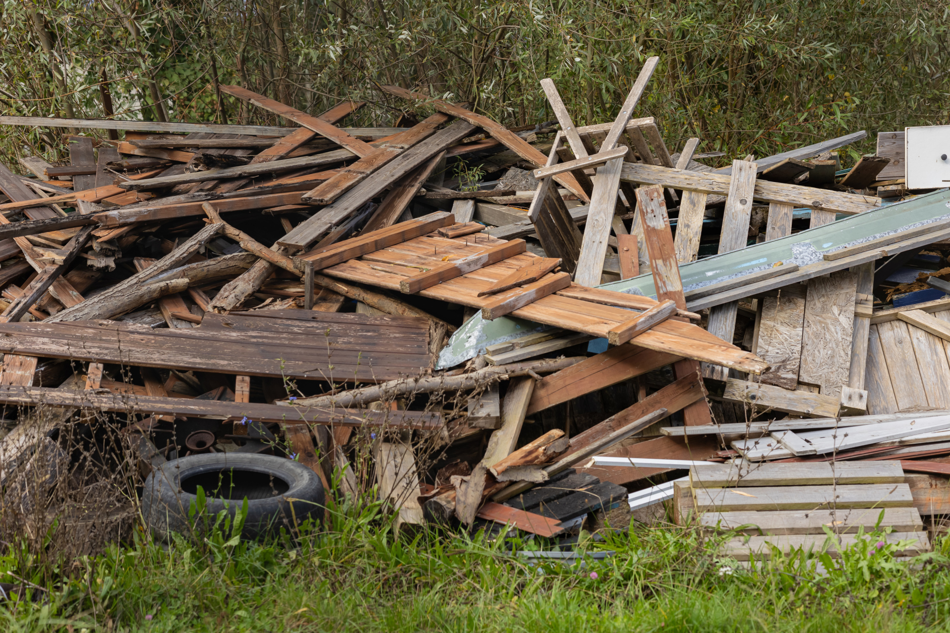 Pile of wooden debris and scrap materials in a rural outdoor area, surrounded by overgrown grass and vegetation. Concept of waste management, recycling, and environmental cleanup Pile of wooden debris and scrap materials in a rural outdoor area, surrounded by overgrown grass and vegetation. Concept of waste management, recycling, and environmental cleanup